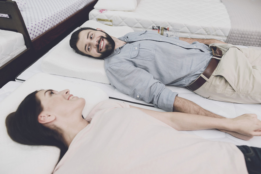 A man in a gray shirt and a woman in a light T-shirt lay on a mattress in the store. They're going to buy a mattress. They lie on it and look at each other