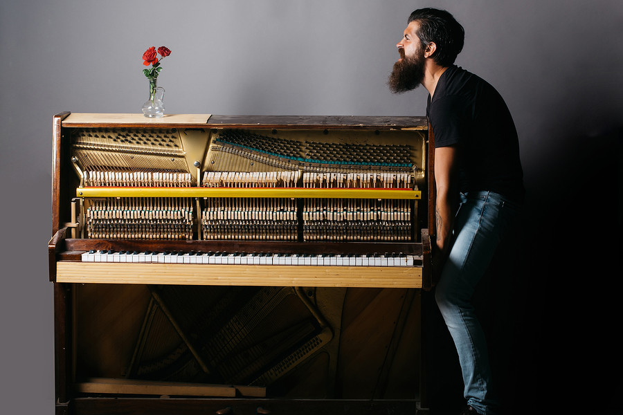 handsome bearded strong man with stylish hair mustache and beard trying to move old wooden or wood open piano with keyboard and glass vase with red rose flowers on grey background copy space