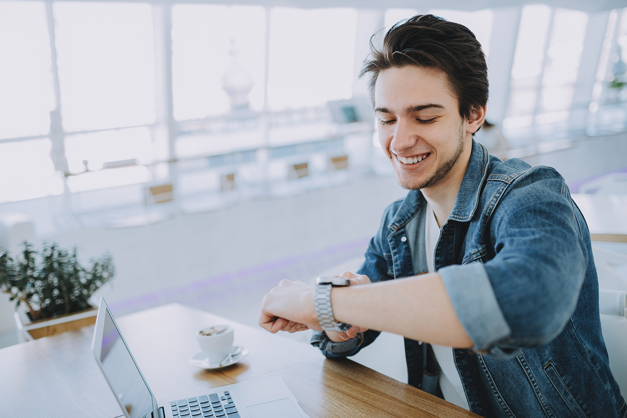 Young stylish man working on a macbook or laptop in cafe while having coffee and checking his nice watch. Happy guy in jeans jacket pointing at clock face