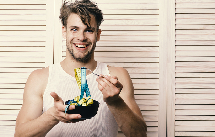 Fitness lifestyle and regime idea. Man with unshaven face holds bowl and fork with measuring tape. Weight management and sportive diet concept. Guy with happy face on background of beige jalousie.