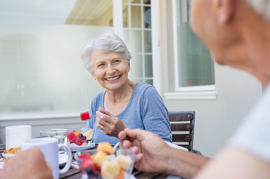 Happy senior woman eating fresh fruits during breakfast. Cheerful old lady with enjoying healthy breakfast with her husband outdoor. Elderly couple in conversation enjoying fruit on the balcony.