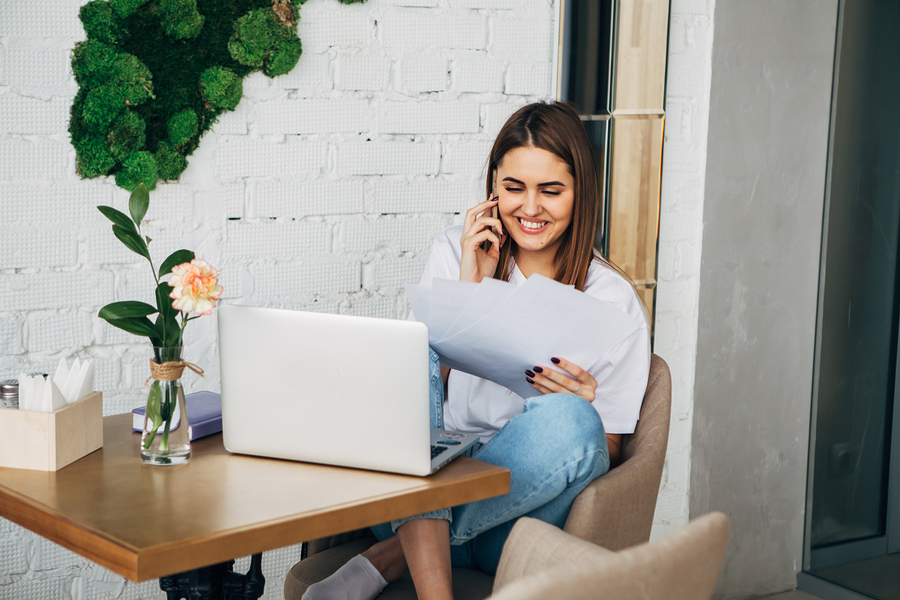 young freelancer girl talking on the phone, holding a notebook, laptop on the table, Beautiful Young Freelancer Woman Using Laptop Computer Sitting At Cafe Table. Happy Smiling Girl Working Online Or Studying And Learning While Using Notebook. Freelance W