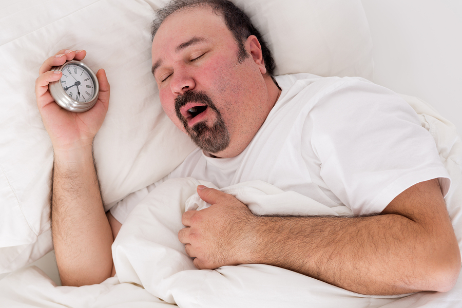 Lethargic tired man lying in bed yawning as he struggles to wake up unmotivated to start the new day and content to rather continue lying in bed as he holds his alarm clock in his hand