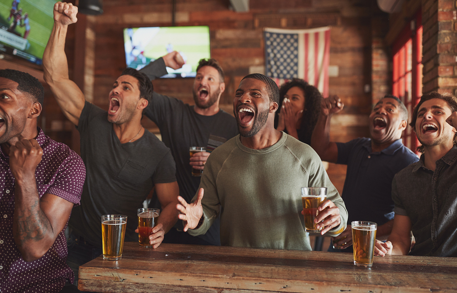 Group Of Male Friends Celebrating Whilst Watching Game On Screen In Sports Bar