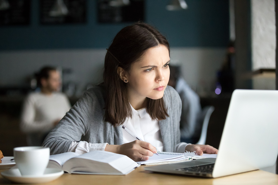 Focused girl concerned about difficult online assignment looking at laptop screen studying working in cafe, millennial student thinking of problem solution reading hard task on computer making notes