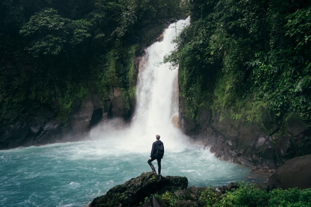 A man outdoors in front of a waterfall enjoying an outdoor travelling trend.